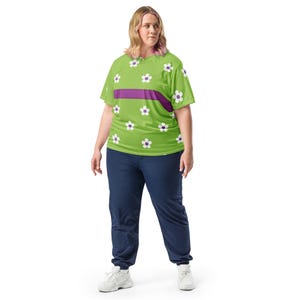 Puede incluir: Una camiseta verde lima con un estampado floral y una franja horizontal morada. La camiseta presenta flores blancas con centros morados. La persona lleva un pantal&oacute;n de ch&aacute;ndal azul marino y zapatillas blancas.