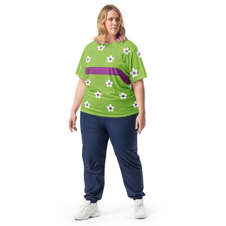 Puede incluir: Una camiseta verde lima con un estampado floral y una franja horizontal morada. La camiseta presenta flores blancas con centros morados. La persona lleva un pantal&oacute;n de ch&aacute;ndal azul marino y zapatillas blancas.