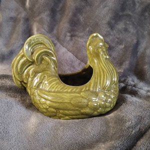 May include: A green ceramic rooster planter. The rooster is in a sitting position with its head turned to the side. The planter has a hole in the top for drainage.