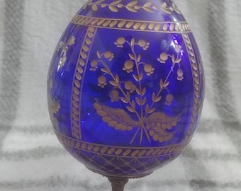 Puede incluir: Un huevo de vidrio azul con diseños florales dorados y un borde dorado, sentado en un soporte de metal dorado.