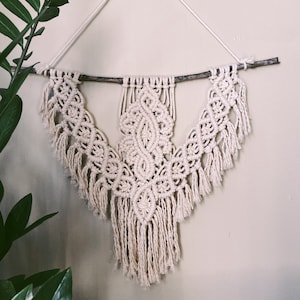 May include: A white macrame wall hanging with a geometric design and fringe. The hanging is attached to a natural branch.