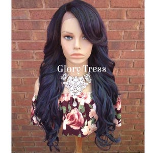 May include: A long, wavy, dark blue wig with teal highlights. The wig is styled with a side part and a side-swept bang. The wig is on a mannequin head.