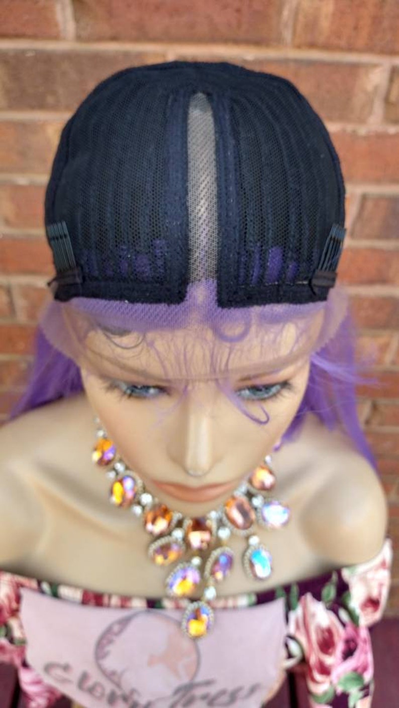 K&ouml;nnte beinhalten: Eine lila Per&uuml;cke mit einem Spitzenfront und verstellbaren Tr&auml;gern. Die Per&uuml;cke sitzt auf einem Mannequin-Kopf mit einem floral gemusterten Hemd und einer Juwelenkette.