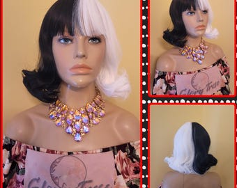 May include: A mannequin head displaying a two-tone wig, black on the left and white on the right, styled in a short, wavy bob. The mannequin wears a large, ornate necklace with colorful gemstones. The off-the-shoulder top has a floral pattern.
