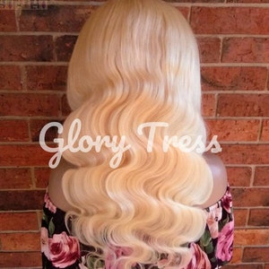 May include: A blonde human hair wig with loose waves. The wig is styled in a long, flowing style. The wig is on a mannequin head and is displayed against a brick wall.