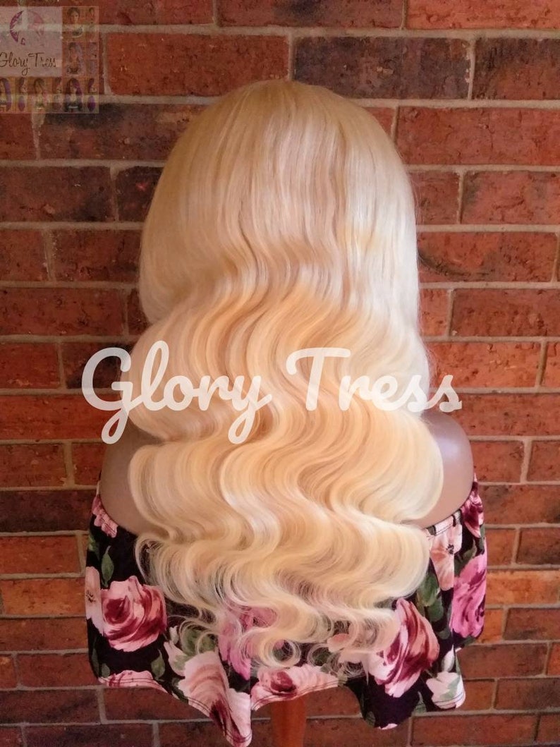 May include: A blonde human hair wig with loose waves. The wig is styled in a long, flowing style. The wig is on a mannequin head and is displayed against a brick wall.