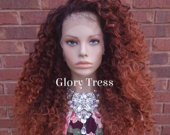 May include: A long, curly, auburn-colored wig with a dark brown root. The wig is styled with a side part and has a lace front. The wig is worn by a mannequin with a floral print top and a large, sparkly necklace. The text "Glory Tress" is visible on the image.