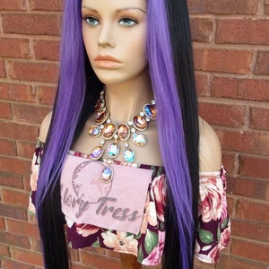 K&ouml;nnte beinhalten: Eine lange, glatte schwarze Per&uuml;cke mit violetten Str&auml;hnen. Die Per&uuml;cke ist mit einem Seitenscheitel gestylt und wird auf einem Mannequin-Kopf getragen. Das Mannequin tr&auml;gt ein Oberteil mit floralem Muster und einen gro&szlig;en, juwelenbesetzten Halsreif.