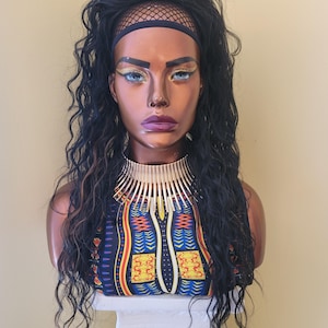 May include: A mannequin head with dark curly hair, wearing a gold collar necklace and a patterned top with blue, yellow, and red accents. The mannequin has dramatic makeup with blue eyeshadow and purple lipstick.