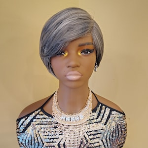 May include: A mannequin head with a short, gray wig and a black and gold sequined top. The mannequin is wearing a gold necklace with a cascading pendant and small dangling earrings. The background is a solid, light yellow color.