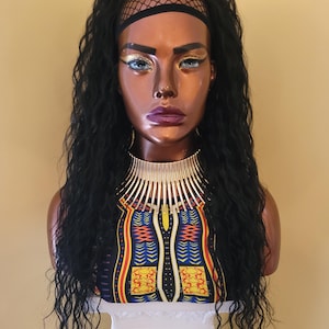 May include: A mannequin head with long, wavy black hair and a black headband. The mannequin wears a gold-toned necklace and a colorful patterned top. The face has dramatic makeup with gold eyeshadow and purple lipstick. The bust is displayed on a white pedestal.