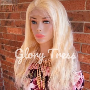 May include: A long, wavy, platinum blonde human hair wig on a mannequin head. The wig has a lace front and is styled in loose waves. The mannequin is wearing a floral print off-the-shoulder top and a gold chain necklace.