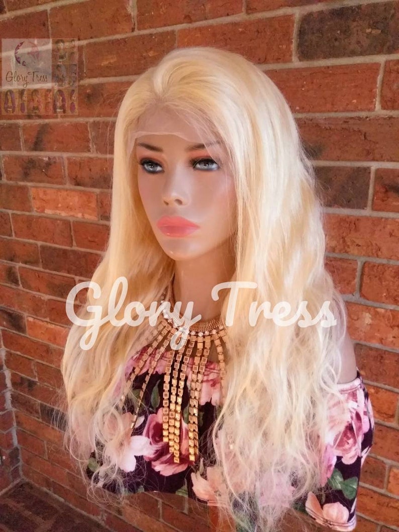 May include: A long, wavy, platinum blonde human hair wig on a mannequin head. The wig has a lace front and is styled in loose waves. The mannequin is wearing a floral print off-the-shoulder top and a gold chain necklace.
