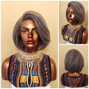 May include: A gray wig with a short, layered style is displayed on a mannequin. The mannequin wears a sleeveless top with a colorful, patterned design in blue, red, and yellow. A beige collar-like accessory is around the neck.