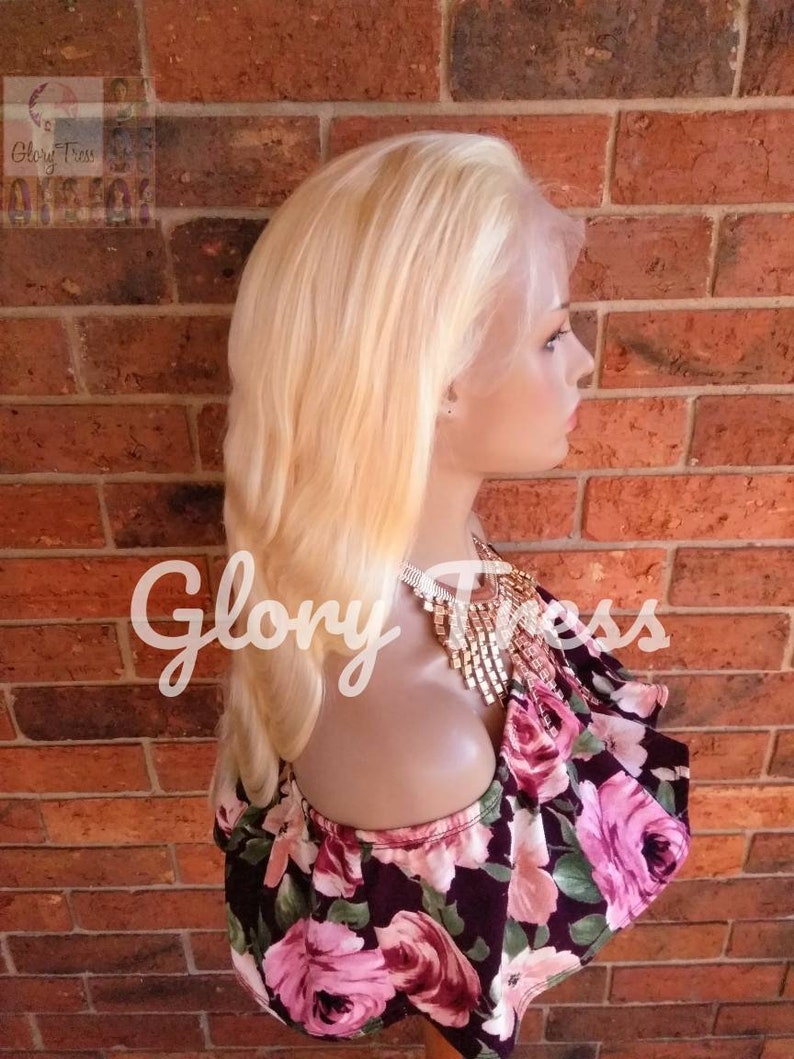 May include: A platinum blonde human hair wig with a lace front. The wig is styled in a straight, shoulder-length cut. The wig is on a mannequin head and is displayed against a brick wall.