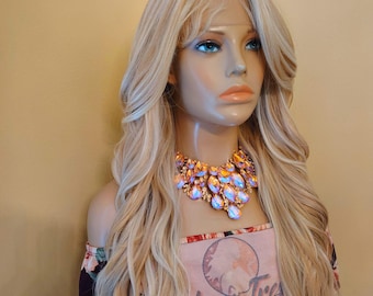 May include: A mannequin head displaying a long, wavy blonde wig with dark roots. The wig is styled with loose curls. The mannequin wears a floral top and a statement necklace with iridescent stones. The text "Glory Tres" is visible on the top.