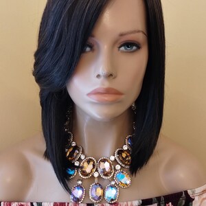 May include: A black bob wig with side-swept bangs. The wig is styled on a mannequin wearing a floral off-the-shoulder top and a large, ornate gold and crystal necklace. The text "Glory Tress" is visible on the top.