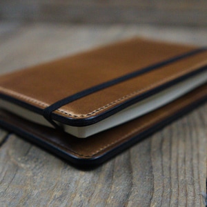 Könnte beinhalten: Ein braunes Lederjournal mit einem schwarzen Gummibandverschluss. Das Journal hat einen genähten Rand und ein schlichtes, elegantes Design. Die Seiten sind weiß und das Journal ist geschlossen. Das Journal liegt auf einer Holzoberfläche.
