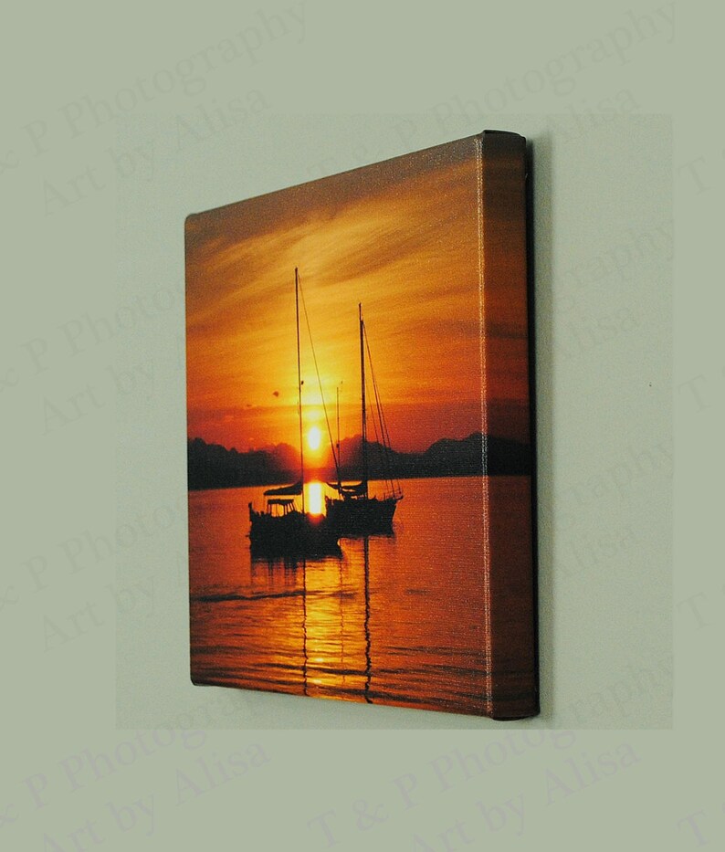 May include: A canvas print of a sunset over the ocean with two sailboats silhouetted against the orange and red sky.