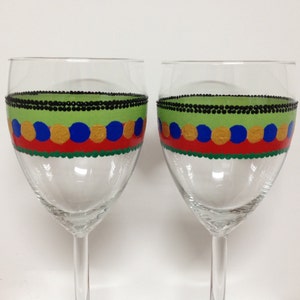 Puede incluir: Dos copas de vino de vidrio transparente con un diseño pintado a mano. El diseño presenta un borde de cuentas negras, una banda verde, una banda roja con círculos dorados y azules, y una banda verde en la parte inferior.