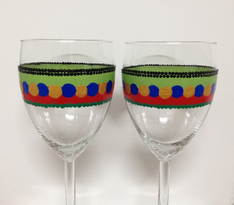 Puede incluir: Dos copas de vino de vidrio transparente con un dise&ntilde;o pintado a mano. El dise&ntilde;o presenta un borde de cuentas negras, una banda verde, una banda roja con c&iacute;rculos dorados y azules, y una banda verde en la parte inferior.