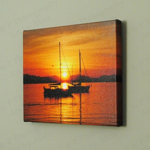 May include: A canvas print of a sunset over the ocean with two sailboats silhouetted against the orange and red sky.