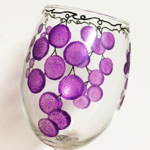 Puede incluir: Una copa de vino de vidrio transparente con un diseño pintado a mano de uvas moradas en una vid. Las uvas están pintadas en un estilo texturizado, dando la apariencia de uvas reales.