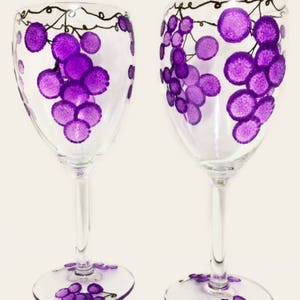 Puede incluir: Dos copas de vino transparentes con racimos de uvas moradas y vides pintadas a mano. La base de cada copa también presenta un diseño de racimo de uvas. Las copas están sobre un fondo claro.