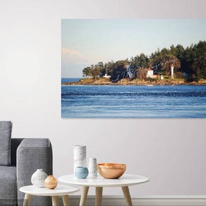 May include: A white lighthouse with a red roof sits on a small island with trees and a blue sky. The water is calm and blue, and there are mountains in the distance.