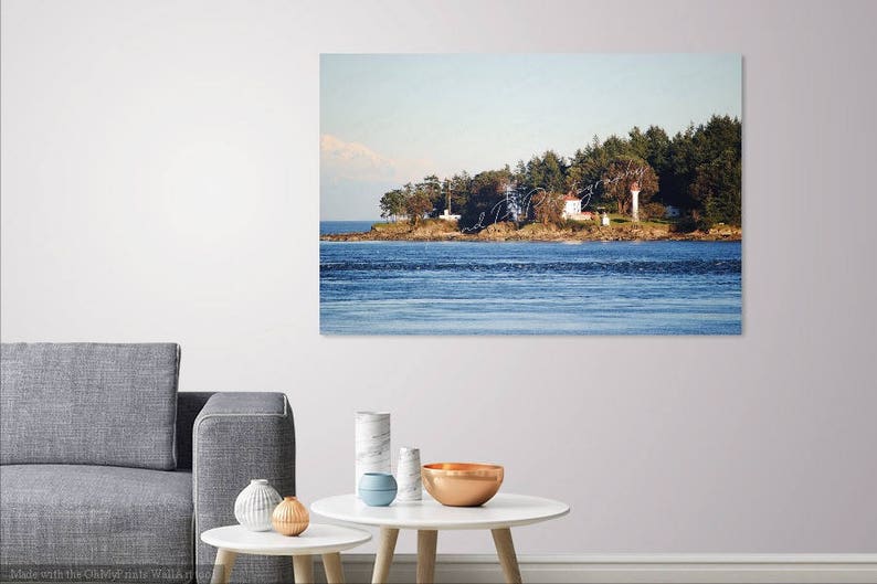 May include: A white lighthouse with a red roof sits on a small island with trees and a blue sky. The water is calm and blue, and there are mountains in the distance.