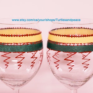 Puede incluir: Dos copas de vino de vidrio transparente con diseños pintados a mano. Las copas presentan un patrón amarillo, verde y rojo con un diseño en zigzag en la parte inferior. El texto "www.etsy.com/ca/your/shops/Turtlesandpeace" es visible en la imagen.