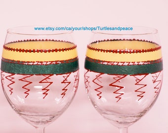 Puede incluir: Dos copas de vino de vidrio transparente con diseños pintados a mano. Las copas presentan un patrón amarillo, verde y rojo con un diseño en zigzag en la parte inferior. El texto "www.etsy.com/ca/your/shops/Turtlesandpeace" es visible en la imagen.