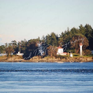 May include: A white lighthouse with a red roof stands on a small island covered in trees. The lighthouse is surrounded by water and a mountain range is visible in the background.