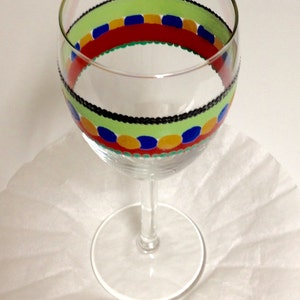 Puede incluir: Una copa de vino de vidrio transparente con un dise&ntilde;o pintado a mano. El dise&ntilde;o presenta una banda verde, negra, roja, azul y amarilla con lunares.