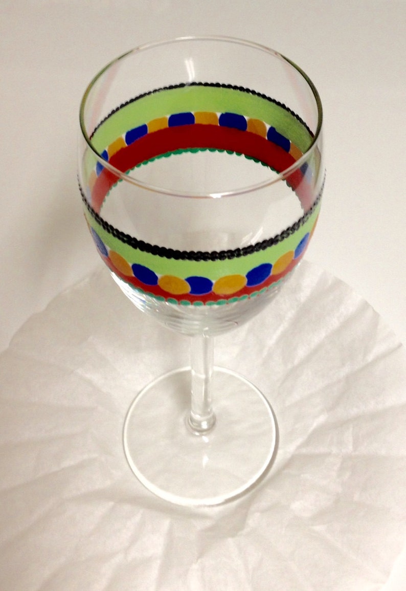 Puede incluir: Una copa de vino de vidrio transparente con un dise&ntilde;o pintado a mano. El dise&ntilde;o presenta una banda verde, negra, roja, azul y amarilla con lunares.