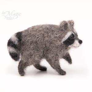 Pode incluir: Uma figura de guaxinim de lã feltrada com um corpo cinzento texturizado, manchas pretas nos olhos e uma cauda às riscas. O guaxinim é representado numa pose de caminhada com um focinho branco. O texto by Magic hand made é visível no canto.