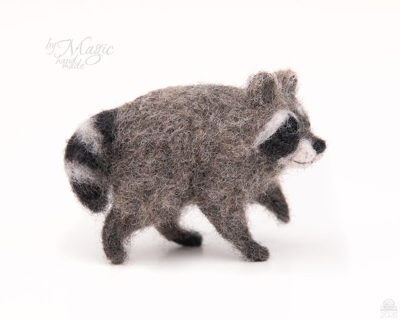 May include: A needle-felted raccoon figurine with a textured gray body, black eye patches, and a striped tail. The raccoon is depicted in a walking pose with a white muzzle and small ears. The text by Magic hand made is visible in the upper left corner.