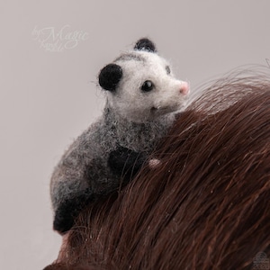 Könnte beinhalten: Eine nadelgefilzte Opossum-Figur mit weißem Gesicht, schwarzen Ohren und einem grauen, strukturierten Körper. Das kleine Tier hat eine rosa Nase und dunkle Augen, mit dem Text by Magic hand made oben links und einem kleinen Datumsstempel 2026.