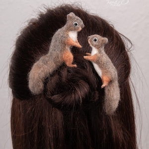 May include: Two needle-felted squirrel hair accessories featuring textured grey and brown fur with white chest patches and dark eyes. The squirrels are positioned upright, one slightly larger than the other, attached to dark brown hair.