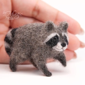May include: A small needle-felted raccoon figurine with textured gray fur, a black and white facial mask, and a striped tail. The raccoon has dark eyes and a small black nose, crafted with a fuzzy, soft appearance in a standing pose.