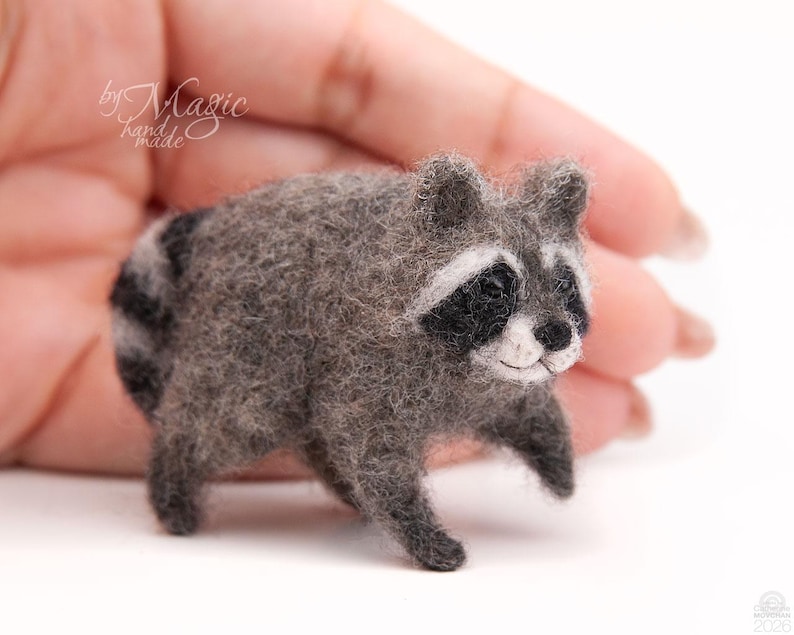 May include: A small needle-felted raccoon figurine with textured gray fur, a black and white facial mask, and a striped tail. The raccoon has dark eyes and a small black nose, crafted with a fuzzy, soft appearance in a standing pose.