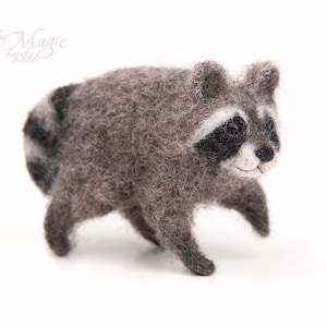 May include: A needle-felted raccoon figurine crafted from gray, black, and white wool fibers. The small animal features a striped tail, dark eye patches, and a textured, fuzzy appearance. The text "by Magic hand made" is visible in the upper left corner.