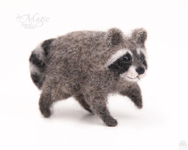May include: A needle-felted raccoon figurine crafted from gray, black, and white wool fibers. The small animal features a striped tail, dark eye patches, and a textured, fuzzy appearance. The text "by Magic hand made" is visible in the upper left corner.