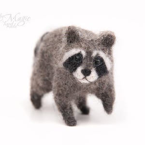 May include: A needle-felted raccoon figurine crafted from fuzzy gray, black, and white wool fibers. The small animal features a distinct black mask around its dark eyes, a white snout, and a textured coat. The text by Magic handmade is visible.