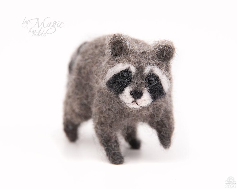 May include: A needle-felted raccoon figurine crafted from fuzzy gray, black, and white wool fibers. The small animal features a distinct black mask around its dark eyes, a white snout, and a textured coat. The text by Magic handmade is visible.