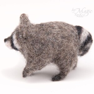 May include: A needle-felted raccoon figurine crafted from textured gray and white wool fibers. The small animal figure features a characteristic black eye mask, pointed ears, and a striped tail. The text "by Magic hand made" is visible in the corner.