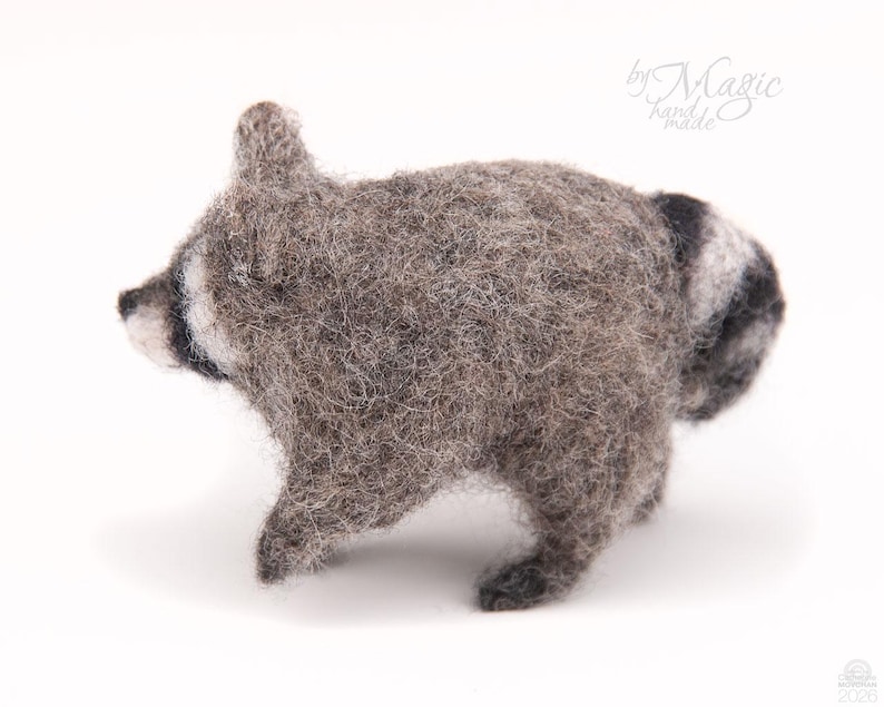 May include: A needle-felted raccoon figurine crafted from textured gray and white wool fibers. The small animal figure features a characteristic black eye mask, pointed ears, and a striped tail. The text "by Magic hand made" is visible in the corner.