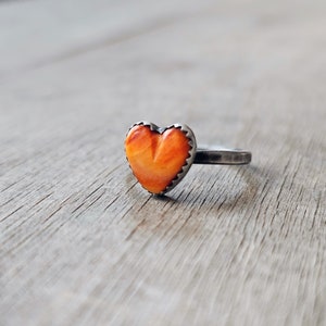 May include: A silver ring featuring an orange heart-shaped stone. The stone has a carved design and is set in a scalloped bezel. The ring band is a simple, textured design.