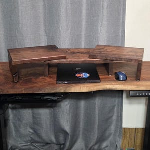 May include: A wooden desk with a live edge, featuring a laptop, a blue mouse, and two wooden monitor stands. The stands are stained a dark brown color. The desk is made of a thick slab of wood.