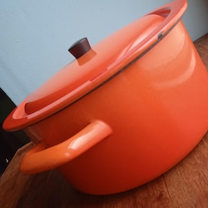 Peut inclure: Une casserole en émail orange avec un couvercle et deux poignées. La casserole est posée sur une surface en bois.